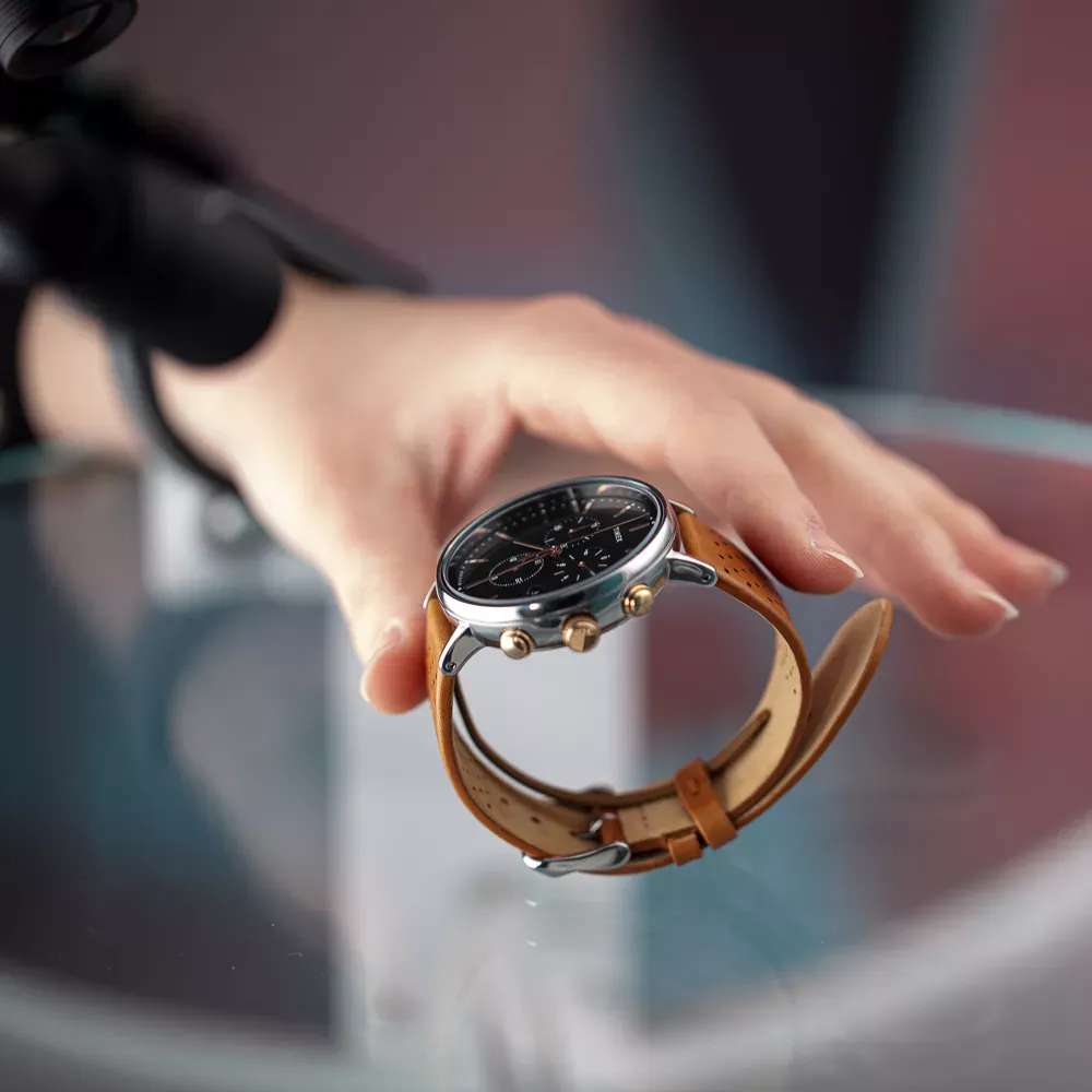 Mano che posiziona orologio da uomo con cinturino in pelle marrone su superficie di vetro
